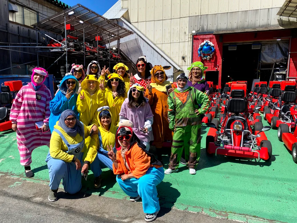 Group of people in colorful costumes posing together near red go-karts on a go-kart track.