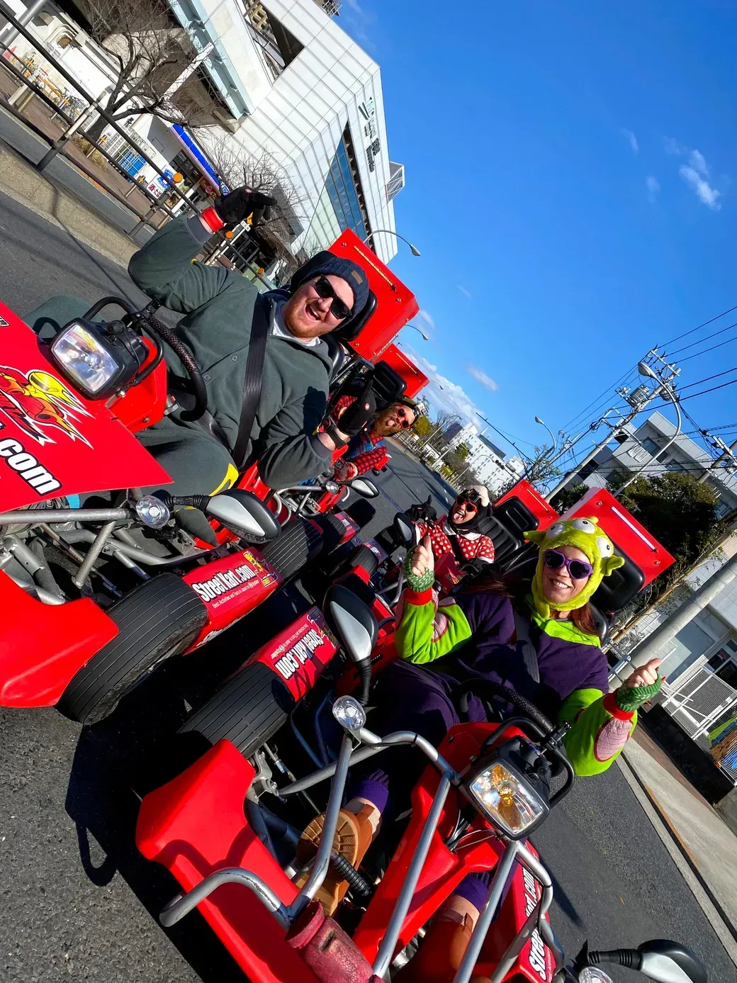 Group of people in colorful costumes posing on bright red go-karts on a city street, smiling and giving thumbs up on a sunny day.