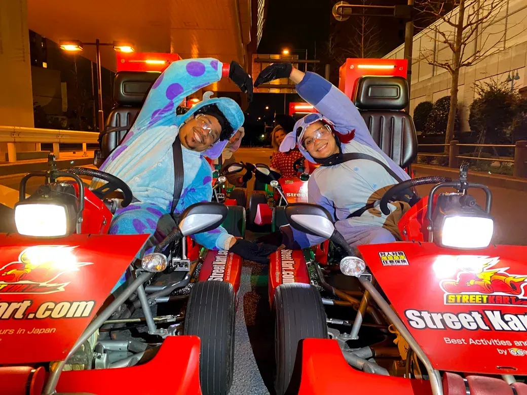 Two people in bright go-karts form a heart with their arms at night on a city street.